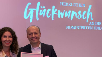Zwei Personen halten eine Urkunde vor einer Leinwand mit "Herzlichen Glückwunsch" beim ATLANTIC Hotel Münster.