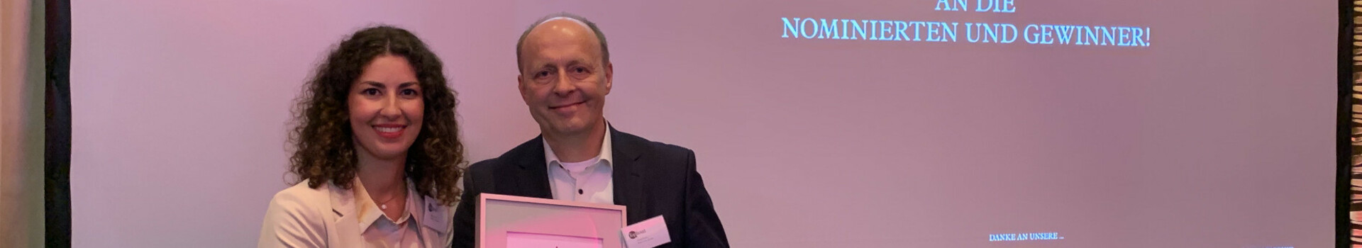 Two people holding a certificate in front of a screen with "Congratulations" at the ATLANTIC Hotel Münster.