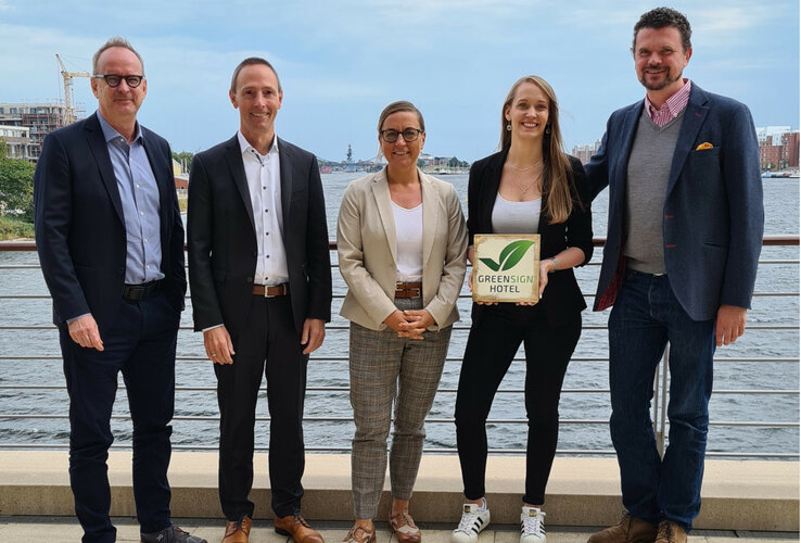 Fünf Personen stehen lächelnd an einem Flussufer, eine hält ein "Greensign Hotel"-Schild.