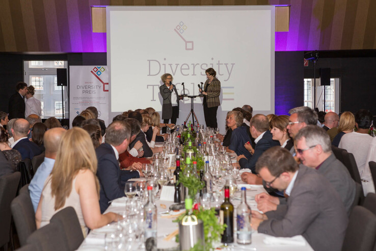 People at a formal dinner with speakers on stage, decorated table with wine glasses and bottles.
