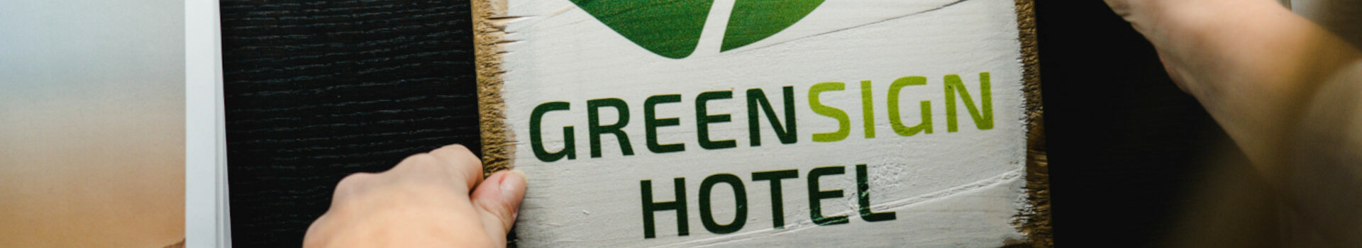 GreenSign Hotel | ATLANTIC Hotel Lübeck ist Level 4 zertifiziert Person hält ein Holzschild mit grünem Blatt und "Greensign Hotel" neben ATLANTIC Hotel Broschüre.