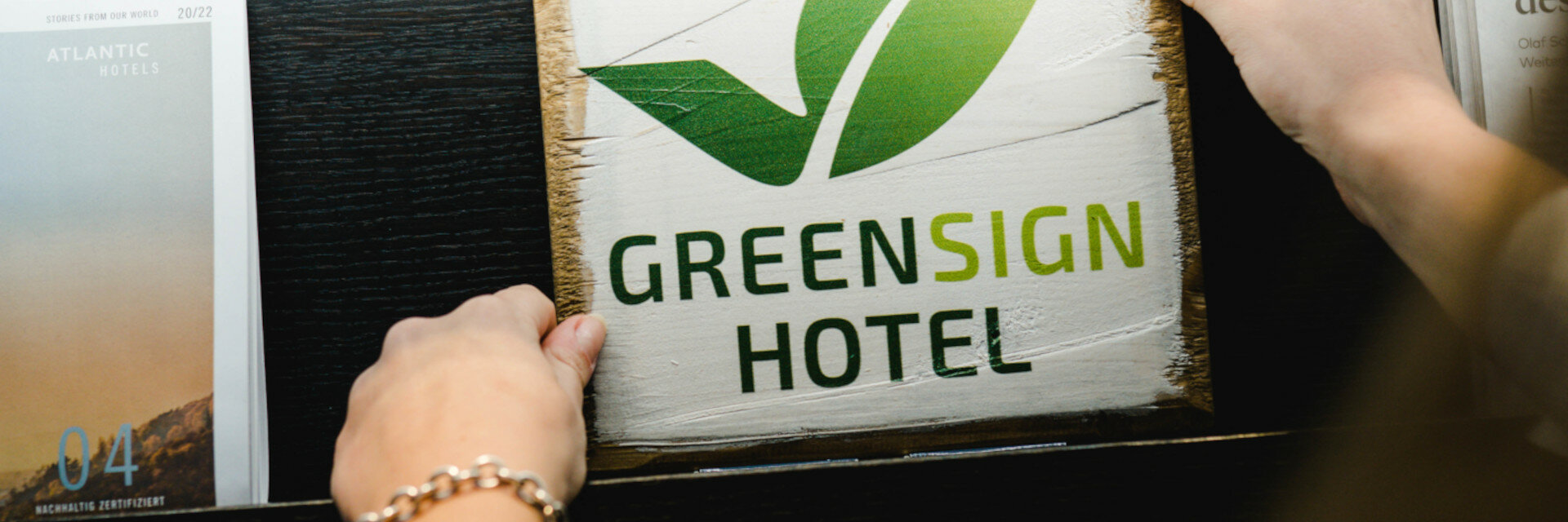 Person hält ein Holzschild mit grünem Blatt und "Greensign Hotel" neben ATLANTIC Hotel Broschüre.