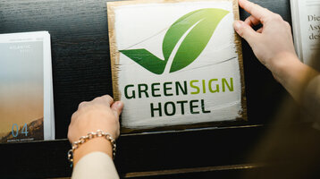 Person holding a wooden sign with a green leaf and "Greensign Hotel" next to ATLANTIC Hotel brochure.