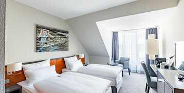 Helles Hotelzimmer mit zwei Einzelbetten, Schreibtisch, Sessel und großem Fenster.