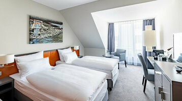 Helles Hotelzimmer mit zwei Einzelbetten, Schreibtisch, Sessel und großem Fenster.