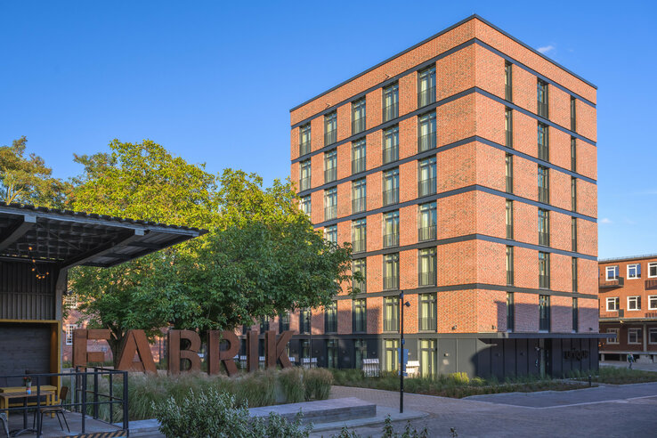 Red brick hotel with several floors, surrounded by trees, under a clear sky.