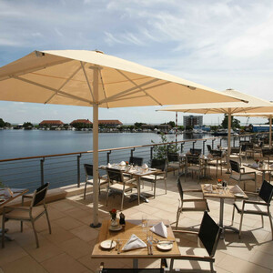 Sonnenterasse im ATLANTIC Hotel Wilhelmshaven Terrasse mit Tischen, Stühlen und Sonnenschirmen, mit Blick auf einen ruhigen See und eine Stadt im Hintergrund.