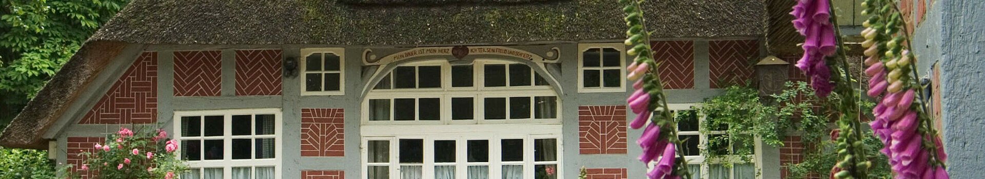 Haus im Schluh | Worpswede Gemütliches Fachwerkhaus mit Reetdach, umgeben von blühenden Pflanzen und Bäumen, mit einer weißen Bank davor.