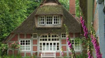 Gemütliches Fachwerkhaus mit Reetdach, umgeben von blühenden Pflanzen und Bäumen, mit einer weißen Bank davor.