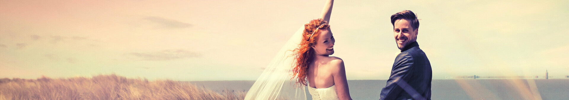 Brautpaar am Strand vor Dünen, die Braut hält einen Blumenstrauß hoch, im Hintergrund das Meer.