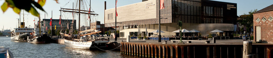 Hafenansicht in Bremerhaven mit Segelschiff und modernem Gebäude am Ufer bei Sonnenuntergang.