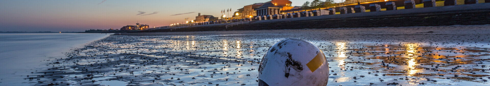 Strand bei Sonnenuntergang mit beleuchteter Promenade und Strandkörben im Hintergrund, Boje im Vordergrund.