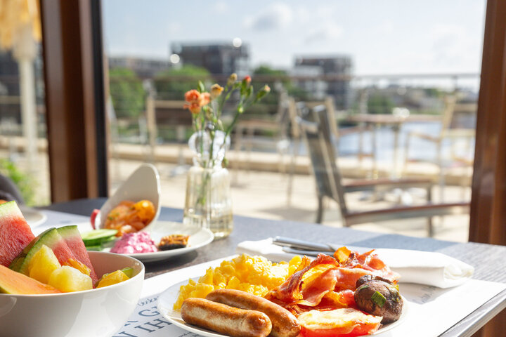 Ein reichhaltiger Frühstücksteller und eine Schale Obst, im Hintergrund die sonnige Außenterrasse