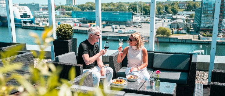 Zwei Gäste sitzen auf der Dachterrasse bei Sonnenschein und stoßen mit Wein an.