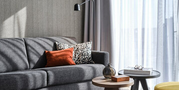 Modernes Hotelzimmer mit grauem Sofa, bunten Kissen, zwei Beistelltischen und großen Fenstern mit Vorhängen.
