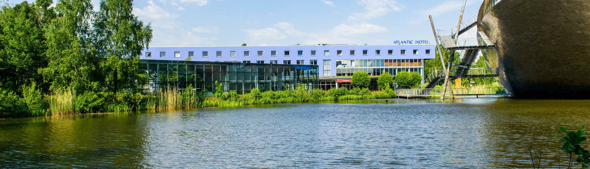 ATLANTIC Hotel Universum am See in Bremen, modernes Gebäude mit Glasfassade, umgeben von Bäumen, blauer Himmel.