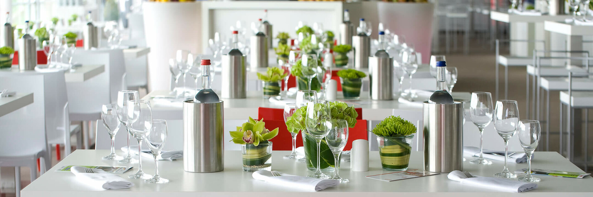 Bright hotel restaurant with elegantly laid tables, flower arrangements and a chef at the buffet in the background.