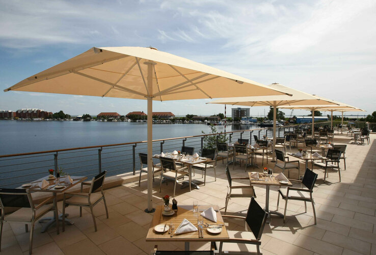 Terrasse eines Restaurants mit Seeblick, Sonnenschirmen und gedeckten Tischen bei klarem Himmel.