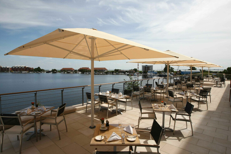 Terrasse eines Restaurants mit Seeblick, Sonnenschirmen und gedeckten Tischen bei klarem Himmel.