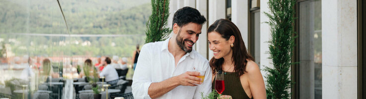 Couple enjoying drinks on the terrace of the ATLANTIC Hotel Heidelberg with a view of green hills.