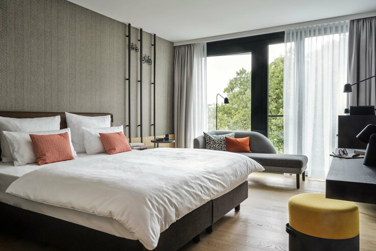 Modern hotel room with double bed, sofa, desk and large windows with a view of the greenery.