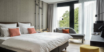 Modernes Hotelzimmer mit Doppelbett, Sofa, Schreibtisch und großen Fenstern mit Blick ins Grüne.