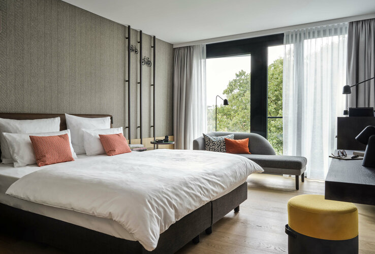 Modernes Hotelzimmer mit Doppelbett, Sofa, Schreibtisch und großen Fenstern mit Blick ins Grüne.