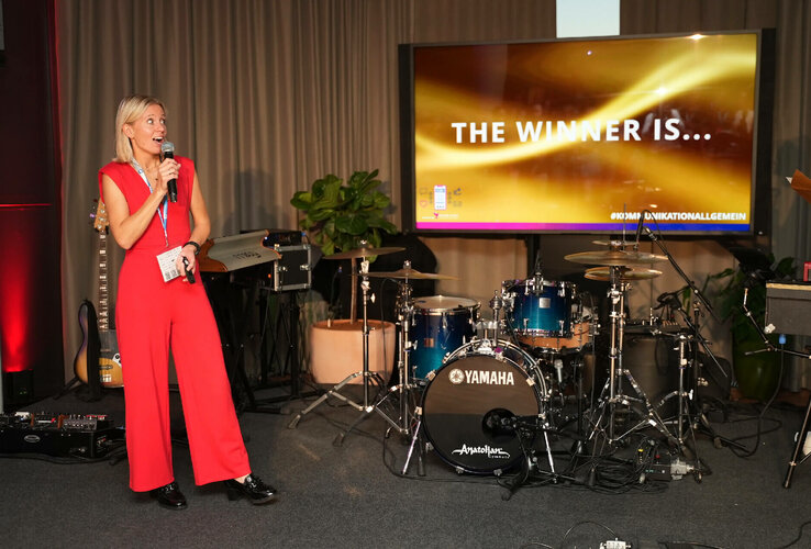 Frau in rotem Anzug spricht in Mikrofon, Bühne mit Schlagzeug, Bildschirm zeigt "The Winner Is..." im ATLANTIC Hotel.