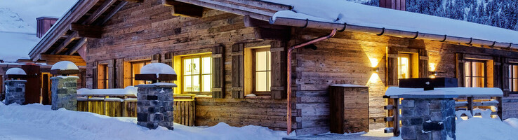 Außenansicht am Abend vom Hotel Severin*s – The Alpine Retreat in Lech Holzhütte im Schnee bei Dämmerung, beleuchtet, mit Berg- und Waldhintergrund.