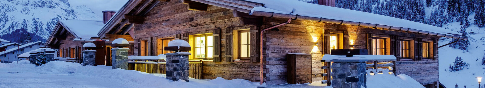 Holzhütte im Schnee bei Dämmerung, beleuchtet, mit Berg- und Waldhintergrund.
