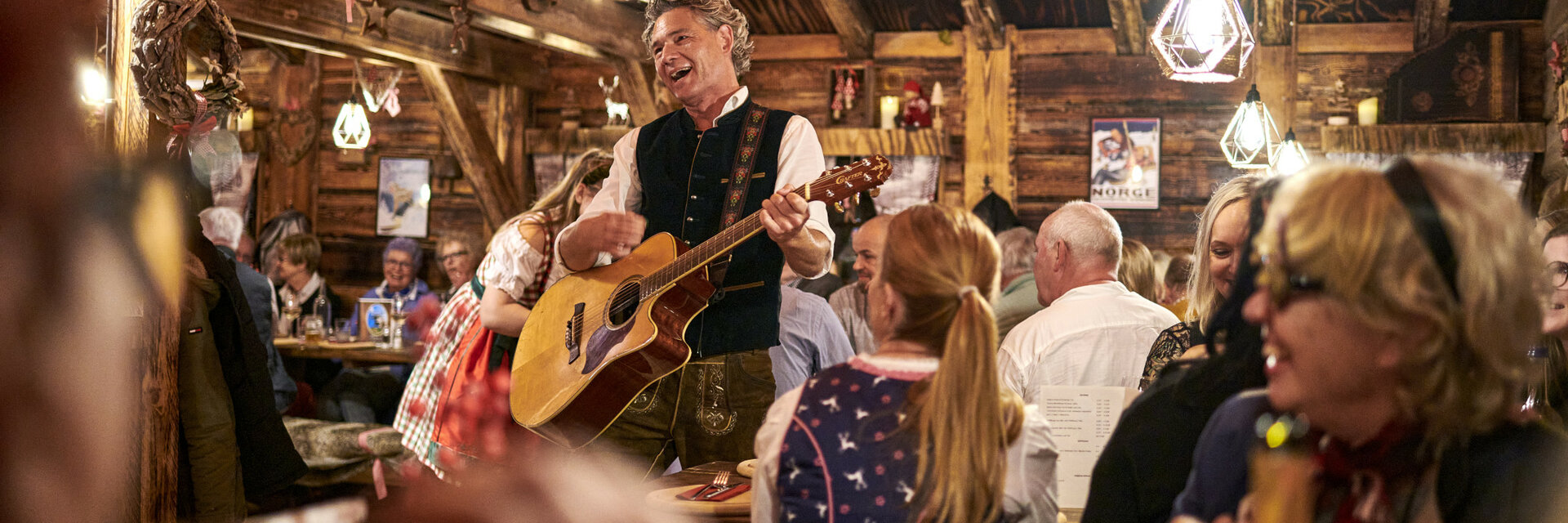 Cozy hut with a musician playing the guitar, surrounded by laughing guests in traditional dress.