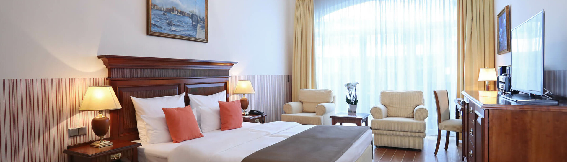 Helles Hotelzimmer mit Doppelbett, Sitzbereich, Kronleuchter, großen Fenstern und Holzmöbeln im ATLANTIC Grand Hotel Travemünde.