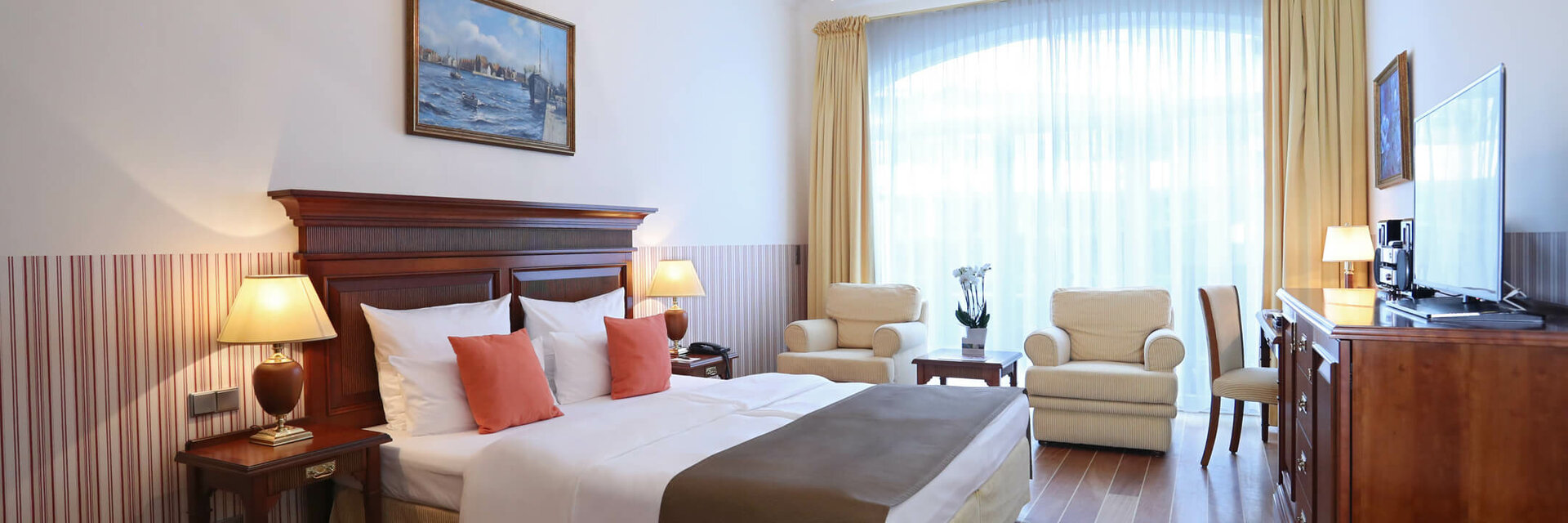 Bright hotel room with double bed, seating area, chandelier, large windows and wooden furniture at the ATLANTIC Grand Hotel Travemünde.