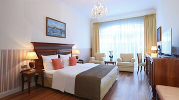 Helles Hotelzimmer mit Doppelbett, Sitzbereich, Kronleuchter, großen Fenstern und Holzmöbeln im ATLANTIC Grand Hotel Travemünde.