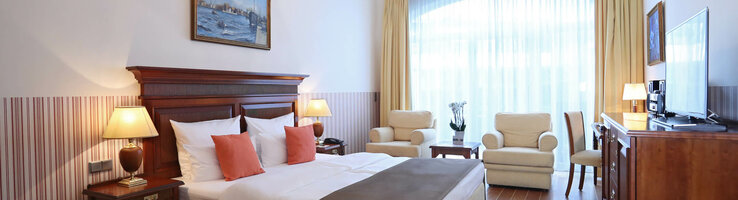 Helles Hotelzimmer mit Doppelbett, Sitzbereich, Kronleuchter, großen Fenstern und Holzmöbeln im ATLANTIC Grand Hotel Travemünde.