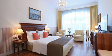 Bright hotel room with double bed, seating area, chandelier, large windows and wooden furniture at the ATLANTIC Grand Hotel Travemünde.
