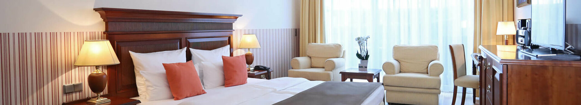 Helles Hotelzimmer mit Doppelbett, Sitzbereich, Kronleuchter, großen Fenstern und Holzmöbeln im ATLANTIC Grand Hotel Travemünde.