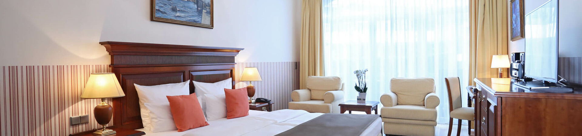 ATLANTIC Grand Hotel Travemünde Zimmer Komfort Helles Hotelzimmer mit Doppelbett, Sitzbereich, Kronleuchter, großen Fenstern und Holzmöbeln im ATLANTIC Grand Hotel Travemünde.