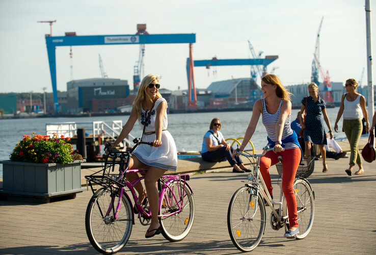 Two women ride along the harbor on bicycles in sunny weather, with cranes and water in the background.
