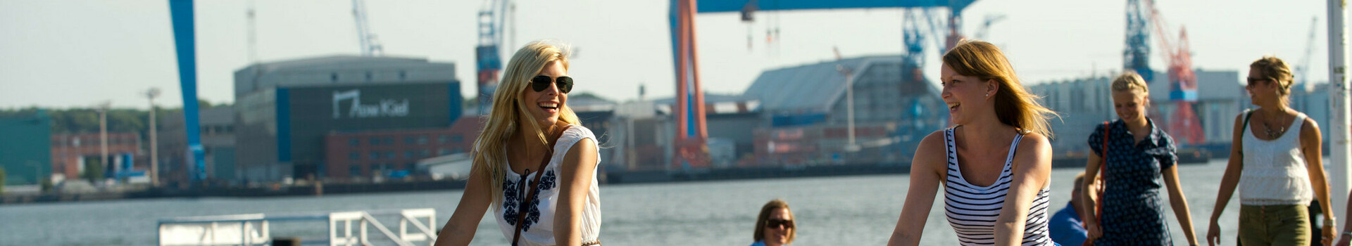Fahrrad fahren in Kiel und Umgebung Zwei Frauen fahren bei sonnigem Wetter am Hafen auf Fahrrädern entlang, im Hintergrund Kräne und Wasser.