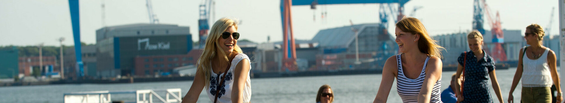 Two women ride along the harbor on bicycles in sunny weather, with cranes and water in the background.