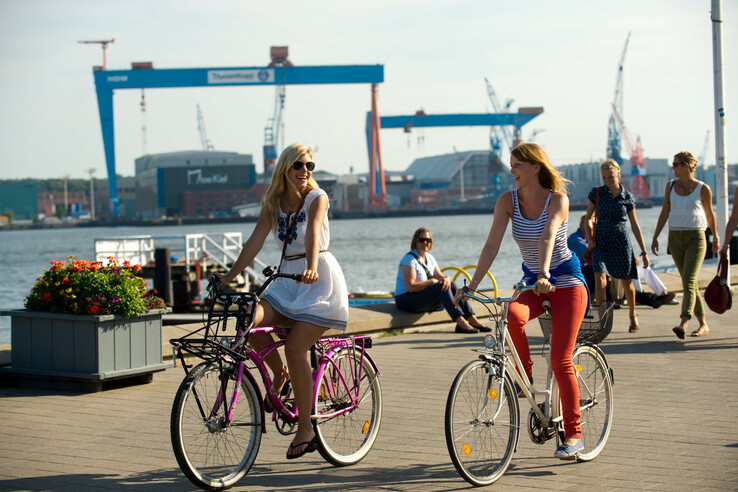 Zwei Frauen fahren bei sonnigem Wetter am Hafen auf Fahrrädern entlang, im Hintergrund Kräne und Wasser.