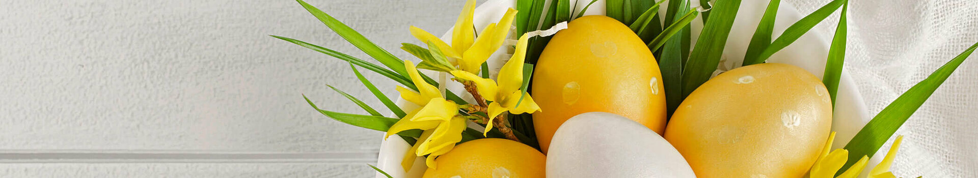 Weiße Schale mit gelben und weißen Ostereiern, dekoriert mit gelbem Blütenschmuck und grünen Blättern auf weißem Tuch.