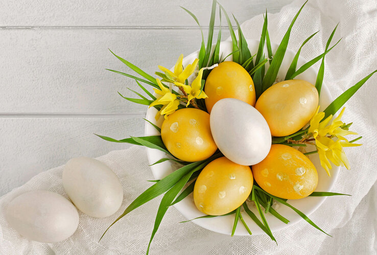 Weiße Schale mit gelben und weißen Ostereiern, dekoriert mit gelbem Blütenschmuck und grünen Blättern auf weißem Tuch.