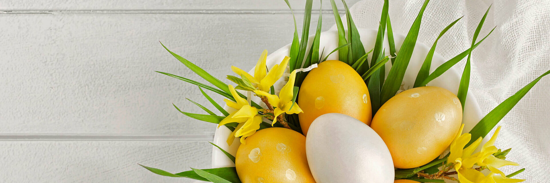 Weiße Schale mit gelben und weißen Ostereiern, dekoriert mit gelbem Blütenschmuck und grünen Blättern auf weißem Tuch.