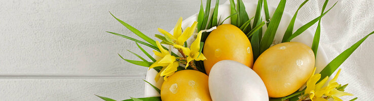 Weiße Schale mit gelben und weißen Ostereiern, dekoriert mit gelbem Blütenschmuck und grünen Blättern auf weißem Tuch.