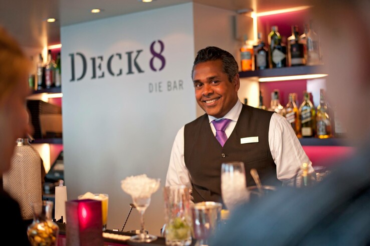 Barkeeper lächelt an der Deck 8 Bar im ATLANTIC Hotel Kiel, umgeben von Getränken und Flaschen.