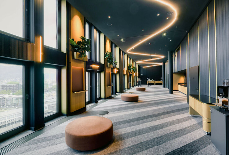 Moderner Hotelflur mit großen Fenstern, geschwungener Beleuchtung, Pflanzen und Sitzgelegenheiten im ATLANTIC Hotel Heidelberg.