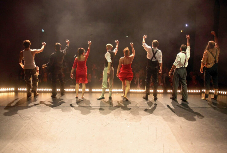 Acht Personen auf einer Bühne, von hinten zu sehen, heben die Arme vor einem applaudierenden Publikum.