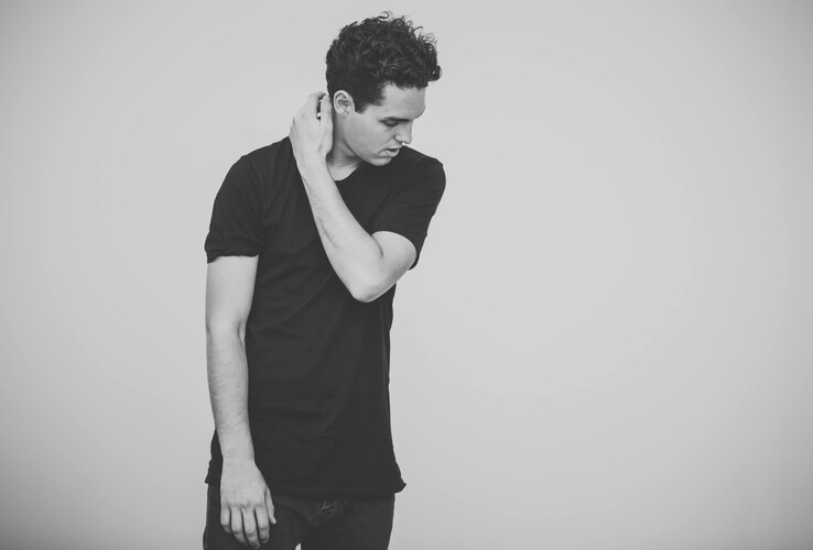 Ein Mann in schwarzem T-Shirt schaut nach unten, vor einem neutralen Hintergrund.